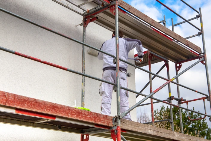 Ouvrier en bâtiment travaillant sur un échafaudage bleu devant une façade de maison en cours de rénovation