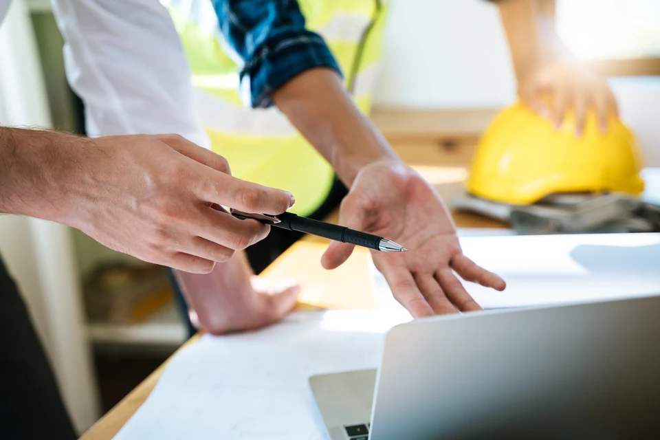Photographie d'un artisan en tenue professionnelle effectuant des travaux de zinguerie sur une toiture en pente, posant ou réparant des éléments de zinc, des gouttières ou des éléments de faîtage, tandis qu'un client particulier observe ou discute avec lui en arrière-plan. La scène se déroule en plein jour sur un chantier résidentiel en Île-de-France. L'image doit illustrer le suivi personnalisé et la relation client tout au long du projet.